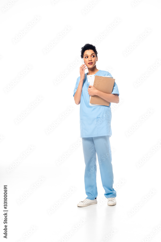 Full length view of african american nurse with papers talking on smartphone on white Full length view of african american nurse with papers talking on smartphone on white