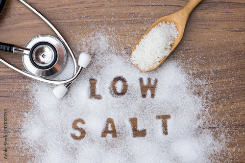 Schilderij op canvas White granulated natural sea salt in wooden scoop and words Low salt  letters written in salt grains and medical stethoscope on wood table background