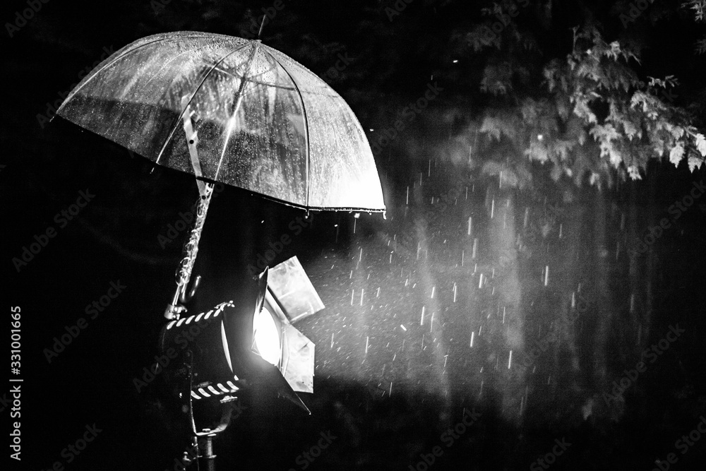 An atmospheric black and white view with shallow depth of field of an umbrella protecting a spotlight from heavy rain during filming video in nature.