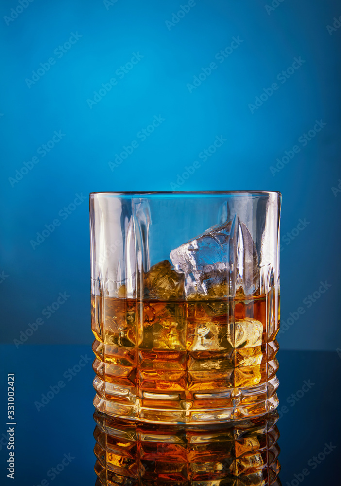 Glass with whiskey and ice on a blue background with a gradient and reflection Glass with whiskey and ice on a blue background with a gradient and reflection