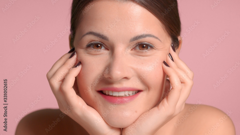 Fototapeta premium Nice pretty brunette with hands support her face watching to the camera and smiling. Pink background, natural makeup