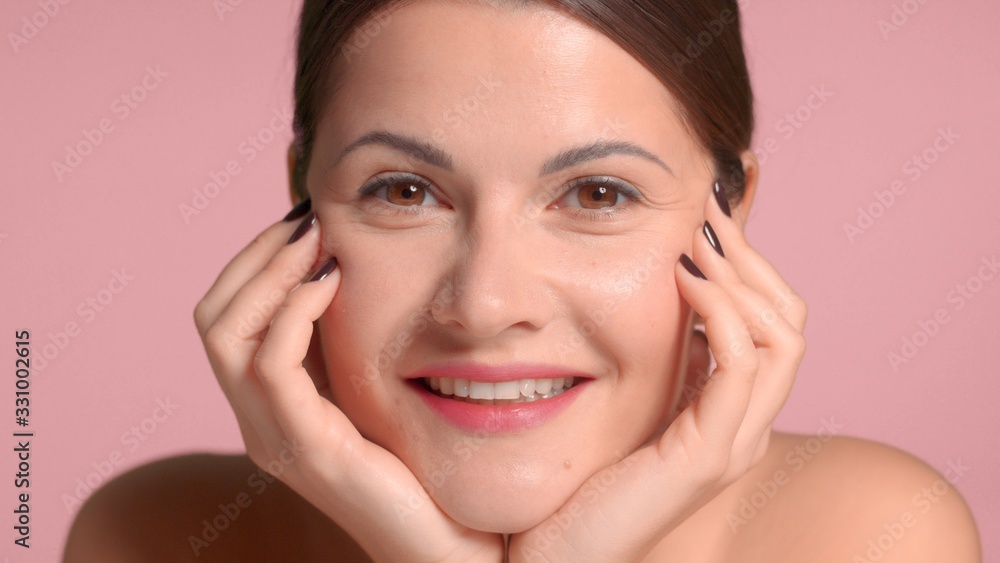 Nice pretty brunette with hands support her face watching to the camera and smiling. Pink background, natural makeup