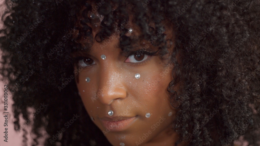 Mixed race black woman with curly hair covered by crystal makeup on pink background in studio Close up show ing makeup