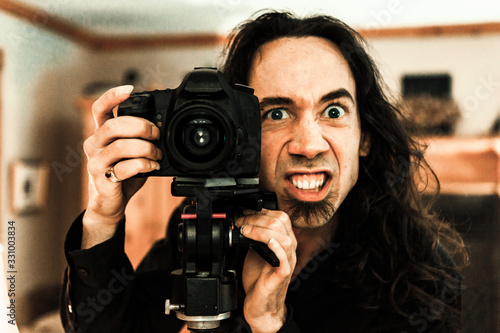 Εκτύπωση καμβά A close up front portrait of a caucasian photographer with a funny creepy face expression, standing behind a professional DSLR camera and tripod