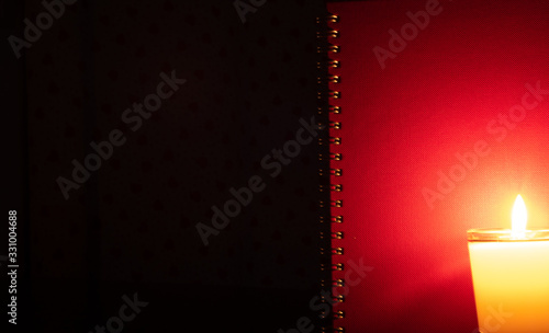 A candle in a burning glass shines in front of the red book cover on a black background.