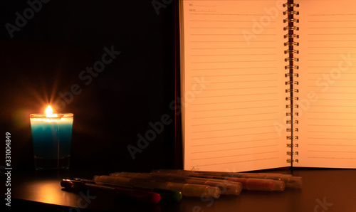 The blue candle in the glass was illuminated on the table beside an open notebook with several pens in the front.