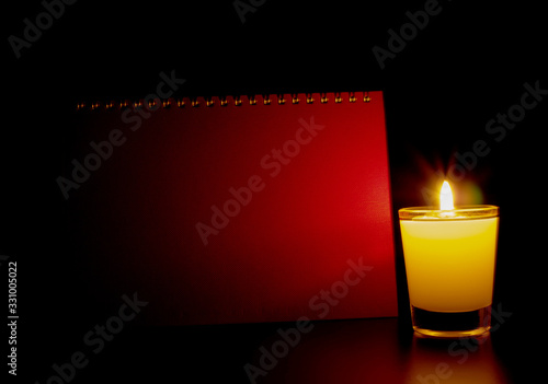A candle in a burning glass shines in front of the red book cover on a black background.