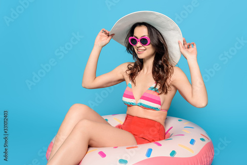 smiling, stylish woman in sunglasses touching sun hat while sitting on swim ring on blue background