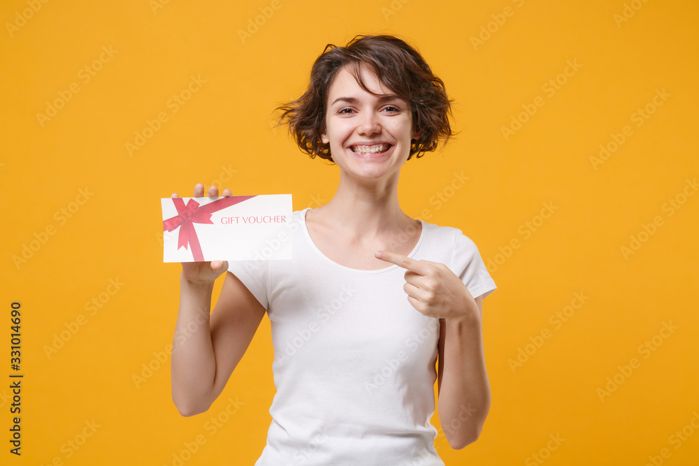 Smiling young brunette woman girl in white t-shirt posing isolated on yellow orange background studio portrait. People lifestyle concept. Mock up copy space. Pointing index finger on gift certificate. Smiling young brunette woman girl in white t-shirt posing isolated on yellow orange background studio portrait. People lifestyle concept. Mock up copy space. Pointing index finger on gift certificate.