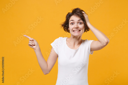 Excited young brunette woman in white t-shirt posing isolated on yellow orange background studio portrait. People lifestyle concept. Mock up copy space. Point index finger aside up, put hand on head.