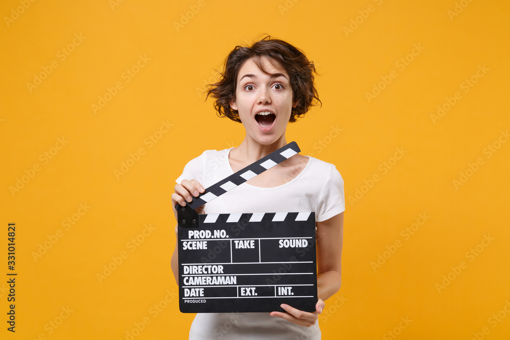 Shocked young brunette woman girl in white t-shirt posing isolated on yellow orange background in studio. People lifestyle concept. Mock up copy space. Hold classic black film making clapperboard.
