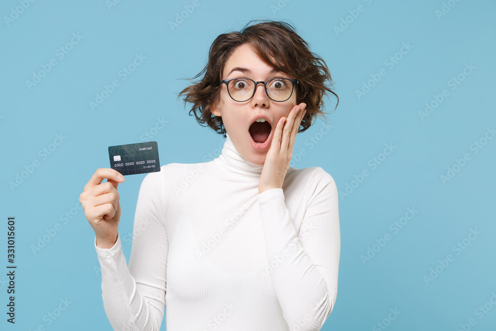 Shocked young brunette woman girl in casual white clothes eyeglasses posing isolated on pastel blue background. People lifestyle concept. Mock up copy space. Hold credit bank card, put hand on cheek. Shocked young brunette woman girl in casual white clothes eyeglasses posing isolated on pastel blue background. People lifestyle concept. Mock up copy space. Hold credit bank card, put hand on cheek.