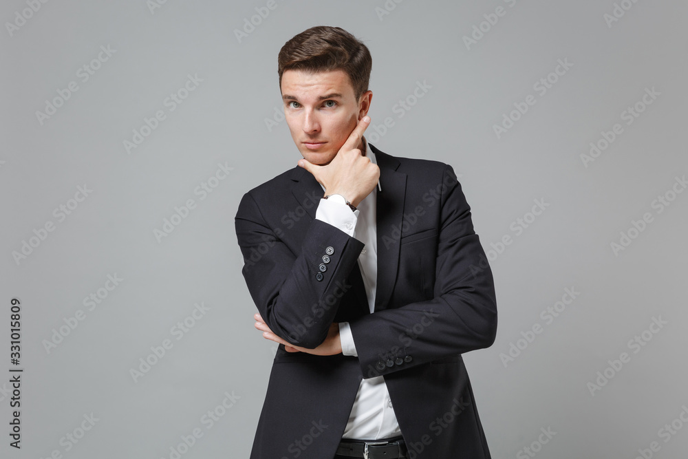 Confident young business man in classic black suit shirt posing isolated on grey background studio portrait. Achievement career wealth business concept. Mock up copy space. Put hand prop up on chin. Confident young business man in classic black suit shirt posing isolated on grey background studio portrait. Achievement career wealth business concept. Mock up copy space. Put hand prop up on chin.