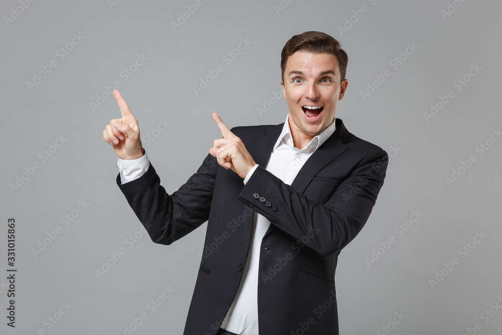 Surprised young business man in classic black suit shirt posing isolated on grey background studio portrait. Achievement career wealth business concept. Mock up copy space. Pointing index fingers up. Surprised young business man in classic black suit shirt posing isolated on grey background studio portrait. Achievement career wealth business concept. Mock up copy space. Pointing index fingers up.