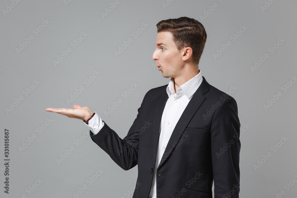 Shocked young business man in classic black suit shirt posing isolated on grey wall background studio portrait. Achievement career wealth business concept. Mock up copy space. Pointing hand aside. Shocked young business man in classic black suit shirt posing isolated on grey wall background studio portrait. Achievement career wealth business concept. Mock up copy space. Pointing hand aside.