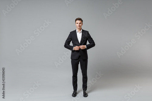 Confident young business man in classic black suit shirt posing isolated on grey background studio portrait. Achievement career wealth business concept. Mock up copy space. Fastening button on jacket.