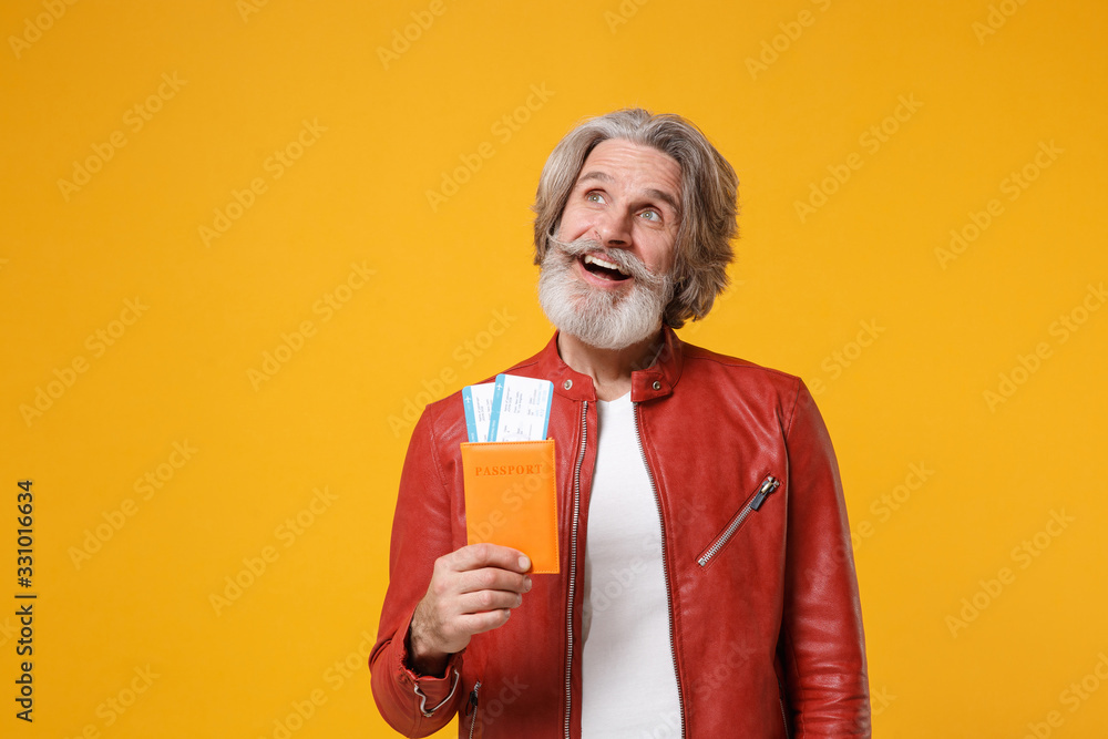 Pensive elderly gray-haired mustache bearded man in red leather jacket isolated on yellow background. People lifestyle concept. Mock up copy space. Hold passport tickets boarding pass, looking up. Pensive elderly gray-haired mustache bearded man in red leather jacket isolated on yellow background. People lifestyle concept. Mock up copy space. Hold passport tickets boarding pass, looking up.