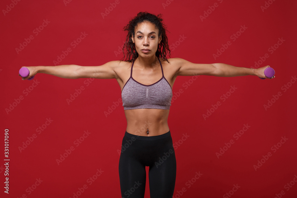 Strong young african american sports fitness woman in sportswear work out isolated on red background. Sport exercises healthy lifestyle concept. Spreading hands, doing exercise for arms with dumbbell. Strong young african american sports fitness woman in sportswear work out isolated on red background. Sport exercises healthy lifestyle concept. Spreading hands, doing exercise for arms with dumbbell.