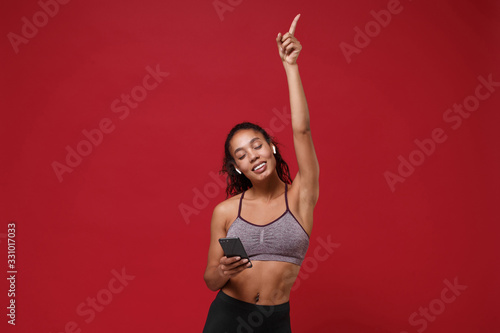 Young african american sports fitness woman in sportswear posing working out isolated on red background. Sport exercises healthy lifestyle concept. Hold mobile phone listen music with earpods dancing.