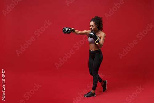 Strong young african american sports fitness boxer woman in sportswear working out isolated on red wall background. Sport exercise healthy lifestyle concept. Doing boxing exercises in boxing gloves.