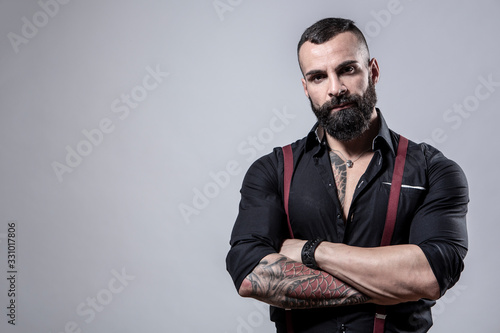 Ragazzo barbuto con Tatuaggi in tutto il corpo, vestito con camicia nera e bretelle rosse,, incorcia le braccia,  isolato su sfondo grigio photo