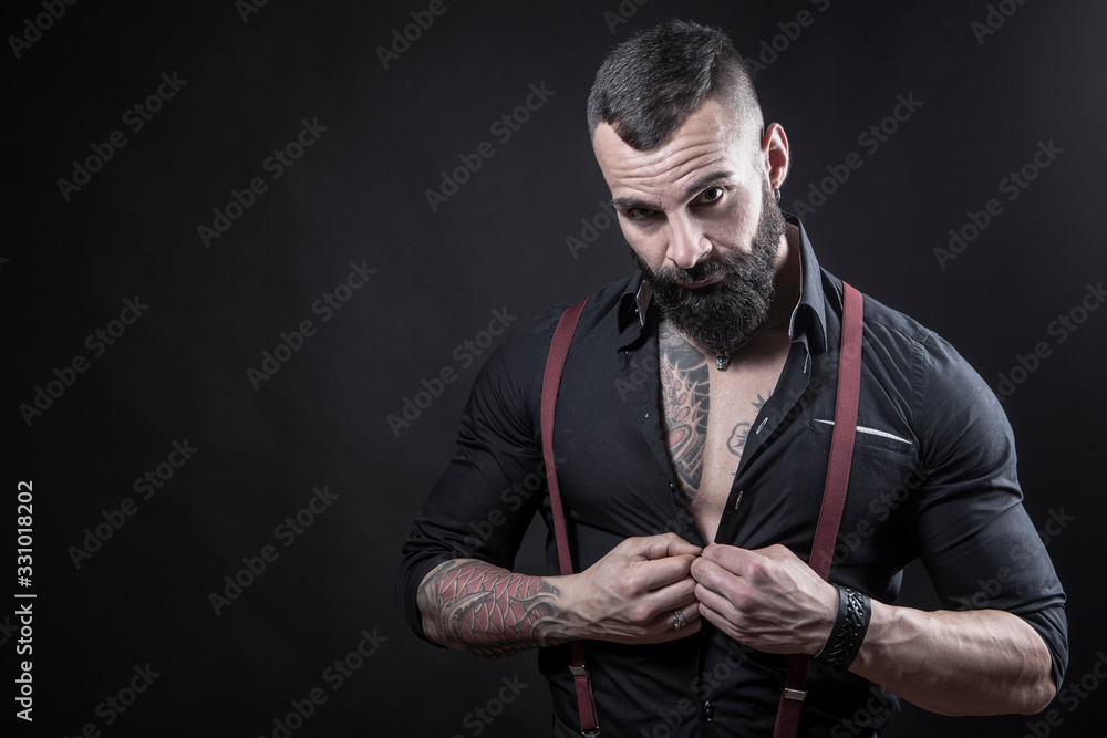 Ragazzo barbuto con Tatuaggi in tutto il corpo, vestito con camicia nera e  bretelle rosse, si aggancia un bottone, isolato su sfondo nero Stock Photo  | Adobe Stock