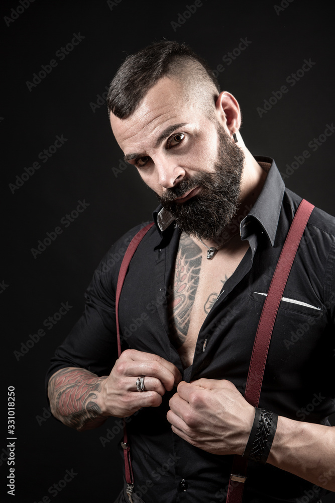 Ragazzo barbuto con Tatuaggi in tutto il corpo, vestito con camicia nera e bretelle  rosse, si aggancia un bottone, isolato su sfondo nero Photos | Adobe Stock