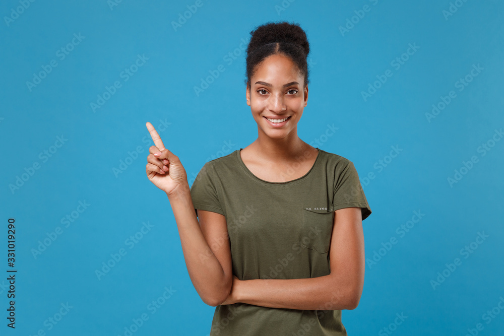 Smiling young african american woman girl in casual t-shirt posing isolated on blue background studio portrait. People sincere emotions lifestyle concept. Mock up copy space. Pointing index finger up. Smiling young african american woman girl in casual t-shirt posing isolated on blue background studio portrait. People sincere emotions lifestyle concept. Mock up copy space. Pointing index finger up.