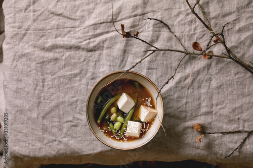 Japanese Miso broth soup with silk tofu cubes, soy beans edamame, green beans in ceramic bowl on grey linen table cloth with autumn branches. Asian dinner. Flat lay, space