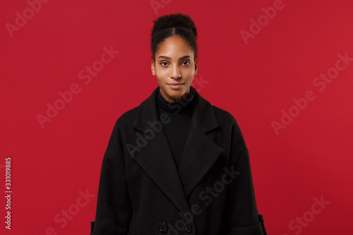 Beautiful young african american woman girl in black coat posing isolated on bright red wall background studio portrait. People sincere emotions lifestyle concept. Mock up copy space. Looking camera.