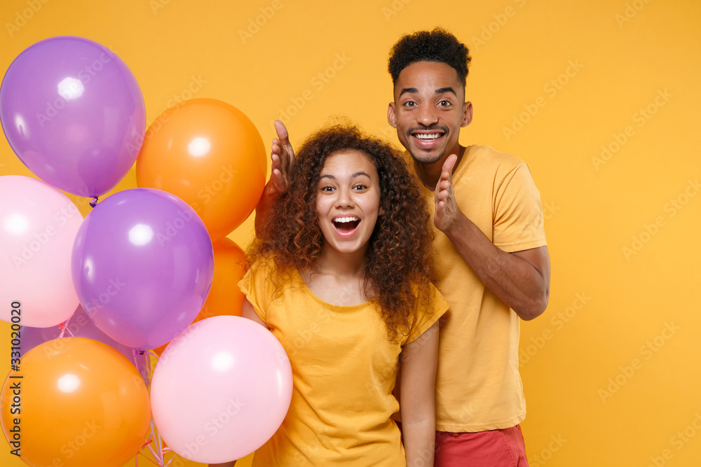 Surprised young friends couple african american guy girl in casual clothes isolated on yellow orange background. Birthday holiday party people emotions concept. Celebrating hold colorful air balloons. Surprised young friends couple african american guy girl in casual clothes isolated on yellow orange background. Birthday holiday party people emotions concept. Celebrating hold colorful air balloons.