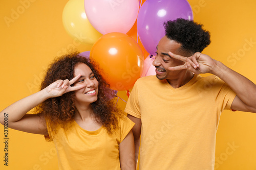 Smiling friends couple african american guy girl in casual clothes isolated on yellow background. Birthday holiday party lifestyle concept. Celebrate hold colorful air balloons showing victory sign.