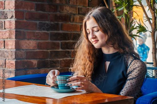 A curly girl with red hair poses in a cafe holding a cup of drink her eyes are directed down