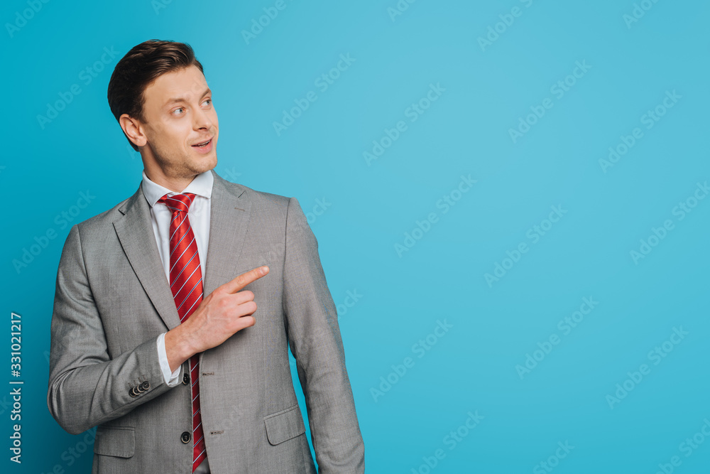 smiling businessman looking away and pointing with finger on blue background smiling businessman looking away and pointing with finger on blue background
