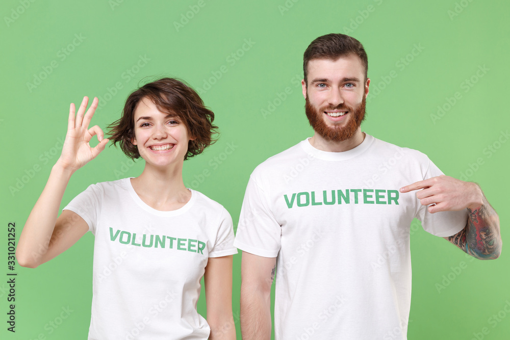 Funny two young friends couple in white volunteer t-shirt isolated on pastel green background studio portrait. Voluntary free work assistance help charity grace teamwork concept. Showing OK gesture. Funny two young friends couple in white volunteer t-shirt isolated on pastel green background studio portrait. Voluntary free work assistance help charity grace teamwork concept. Showing OK gesture.