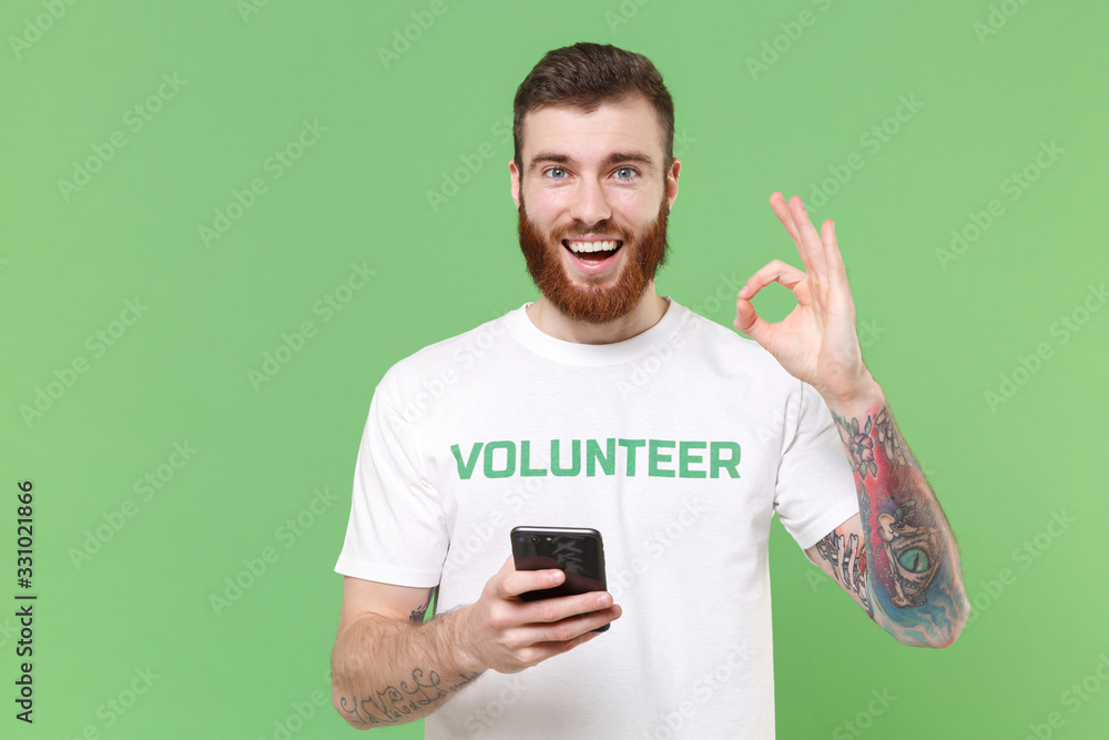 Cheerful young bearded man in white volunteer t-shirt isolated on pastel green background. Voluntary free work assistance help charity grace teamwork concept. Using mobile phone, showing OK gesture. Cheerful young bearded man in white volunteer t-shirt isolated on pastel green background. Voluntary free work assistance help charity grace teamwork concept. Using mobile phone, showing OK gesture.