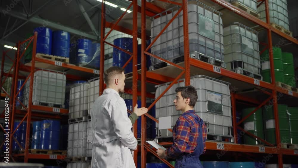 Rear view tracking shot of engineer in white coat explaining task to young warehouse worker standing by steel racks with containers and barrels in industrial storage Rear view tracking shot of engineer in white coat explaining task to young warehouse worker standing by steel racks with containers and barrels in industrial storage