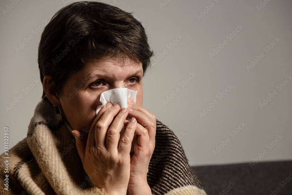 A sick older woman sneezes into a napkin, and covered in a blanket at home. An ill woman sitting on the sofa alone. Coronavirus. COVID-19. A quarantine at home. A sick older woman sneezes into a napkin, and covered in a blanket at home. An ill woman sitting on the sofa alone. Coronavirus. COVID-19. A quarantine at home.