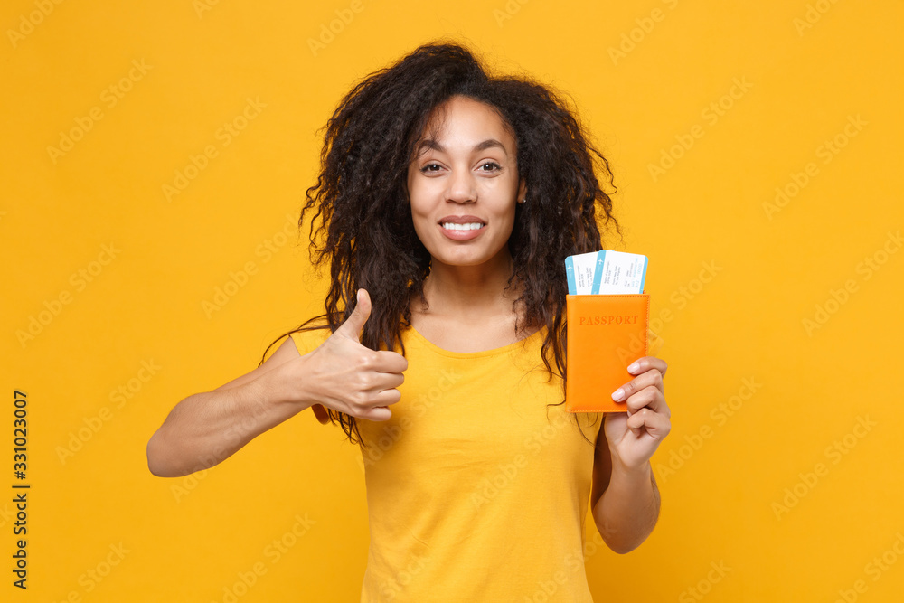 Smiling young african american woman girl in casual t-shirt posing isolated on yellow background. People lifestyle concept. Mock up copy space. Hold passport boarding pass tickets showing thumb up. Smiling young african american woman girl in casual t-shirt posing isolated on yellow background. People lifestyle concept. Mock up copy space. Hold passport boarding pass tickets showing thumb up.
