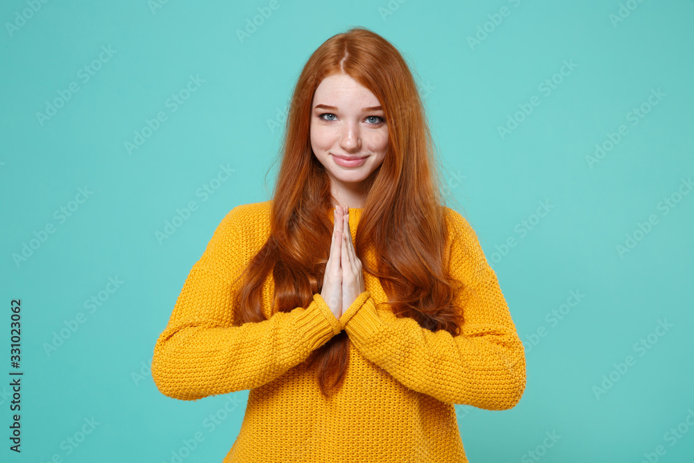 Fototapeta premium Smiling young redhead woman girl in yellow sweater posing isolated on blue turquoise background studio portrait. People emotions lifestyle concept. Mock up copy space. Holding hands folded in prayer.