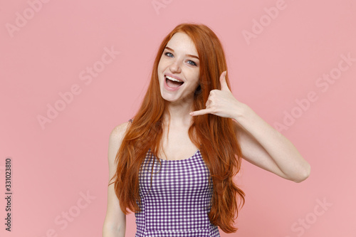 Cheerful young redhead woman girl in plaid dress posing isolated on pastel pink background studio portrait. People lifestyle concept. Mock up copy space. Doing phone gesture like says call me back.