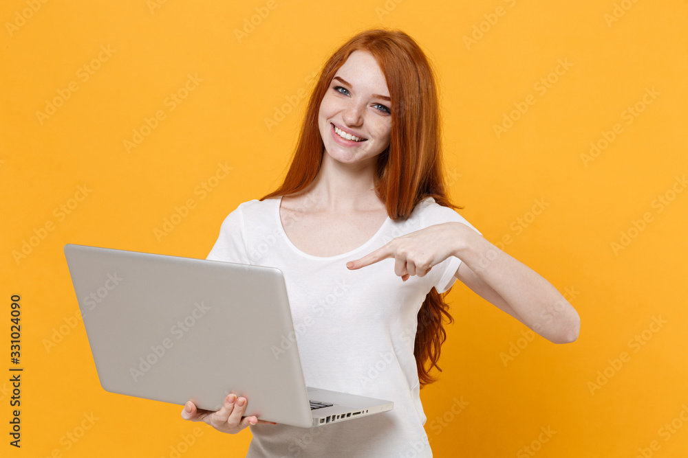 Smiling young redhead woman girl in white blank empty t-shirt posing isolated on yellow background in studio. People lifestyle concept. Mock up copy space. Pointing index finger on laptop pc computer. Smiling young redhead woman girl in white blank empty t-shirt posing isolated on yellow background in studio. People lifestyle concept. Mock up copy space. Pointing index finger on laptop pc computer.