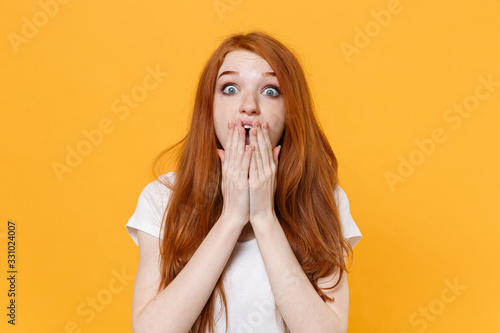 Shocked young redhead woman girl in white blank empty t-shirt posing isolated on yellow background studio portrait. People emotions lifestyle concept. Mock up copy space. Covering mouth with hands.