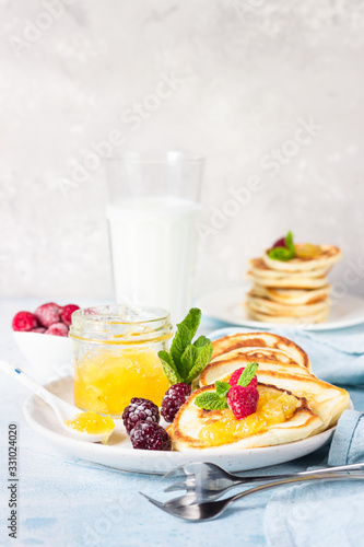 Pancakes with orange jam, frozen berries (raspberries and blackberries), mint and glass with milk on light blue stone background. Idea for children’s breakfast or snack. 