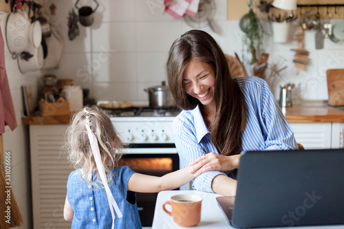 Working mom works from home with kid. Happy mother and daughter have fun. Successful woman and cute child using laptop. Freelancer workplace in kitchen. Female business. Lifestyle authentic moment