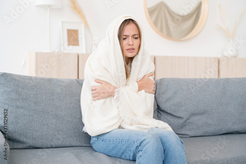 Fotografie Upset young woman freezing, feeling cold at home, sitting on sofa covered with b