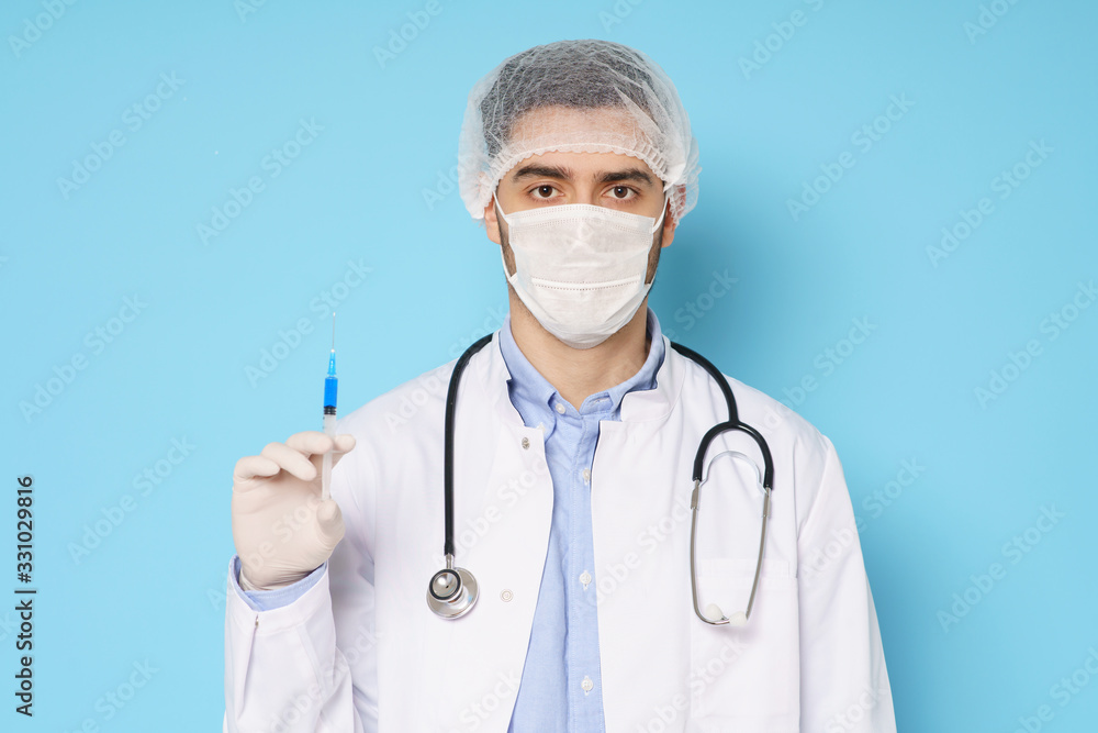 Young doctor wearing white medical mask and gloves holding syringe with coronavirus vaccine, isolated on blue background Young doctor wearing white medical mask and gloves holding syringe with coronavirus vaccine, isolated on blue background