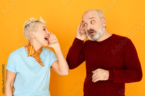 Marriage, relationships, people and age concept. Attractive mature woman with short gray hair screaming while addressing to her husband who is holding hand at his ear because of hearing problem photo