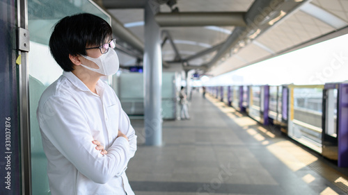 Asian man wearing face mask at skytrain station or urban train platform. Wuhan coronavirus (COVID-19) outbreak prevention in public transportation. Health awareness for PM2.5 air pollution protection.