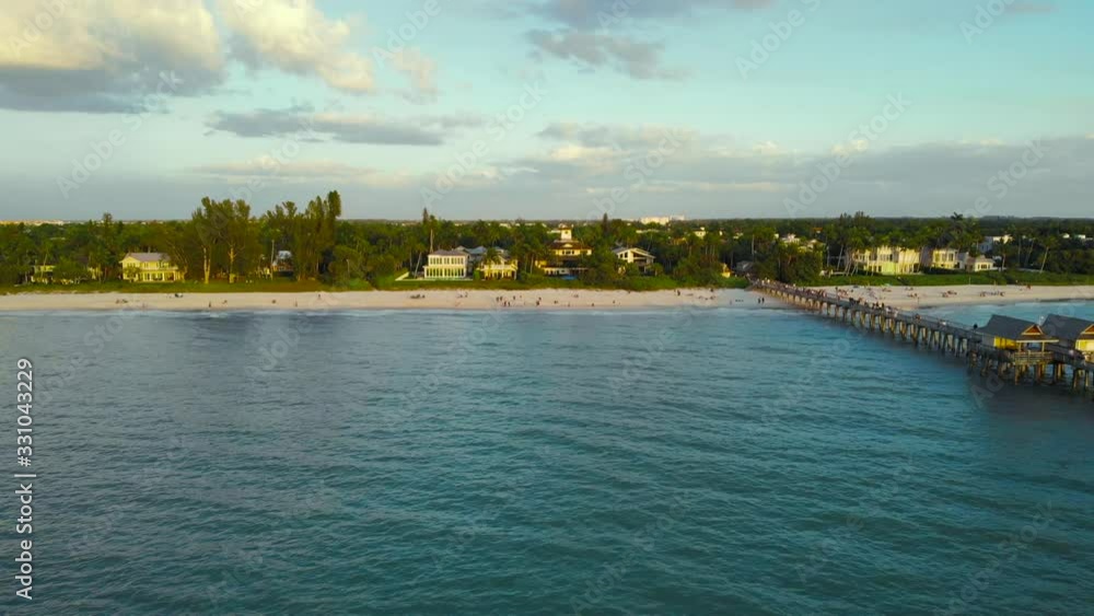 Coast and the beach near the pier leaving into the ocean. Drone flies forward from the ocean to shore along a large wooden fishing pier. People walk along the ocean. Luxury houses near the ocean. Coast and the beach near the pier leaving into the ocean. Drone flies forward from the ocean to shore along a large wooden fishing pier. People walk along the ocean. Luxury houses near the ocean.